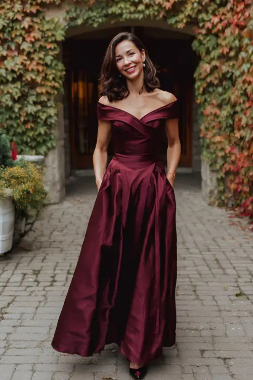 a woman over 50 in a burgundy satin gown with an asymmetrical off-shoulder neckline and pleated A-line skirt