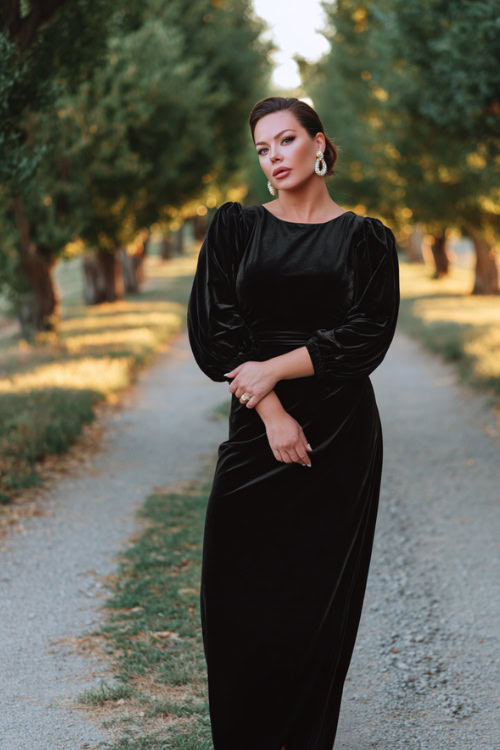 A plus-size woman wears a sleek black velvet gown with long bishop sleeves and a defined waistline, styled with pearl earrings and heels