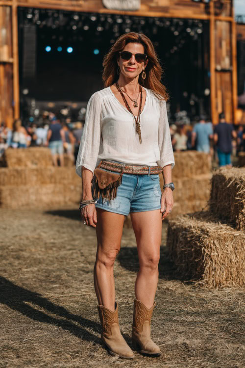 A woman over 40 in denim shorts, a tucked-in lightweight long-sleeve blouse, and tan ankle boots, accessorized with a belt bag and layered necklaces
