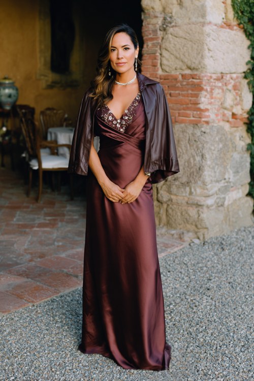 a woman over 30 wears a dark wine satin wrap gown with beadwork at the neckline and flowing cape sleeves, styled with a sleek chocolate leather jacket and pearl studs