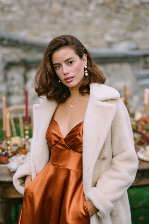 A woman wears a copper satin dress with a cream wool coat and pearl earrings