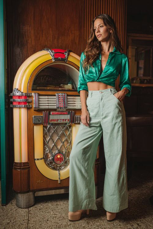 A woman in wide-leg jeans, a silky cropped blouse in emerald green, and platform heels, standing near a jukebox inside a casual fall bar