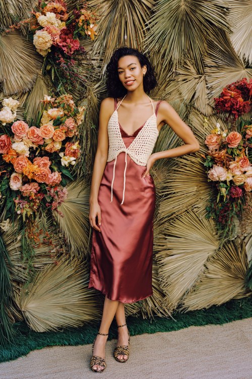 A woman wears a rosewood satin slip midi topped with a cream macrameÌ vest