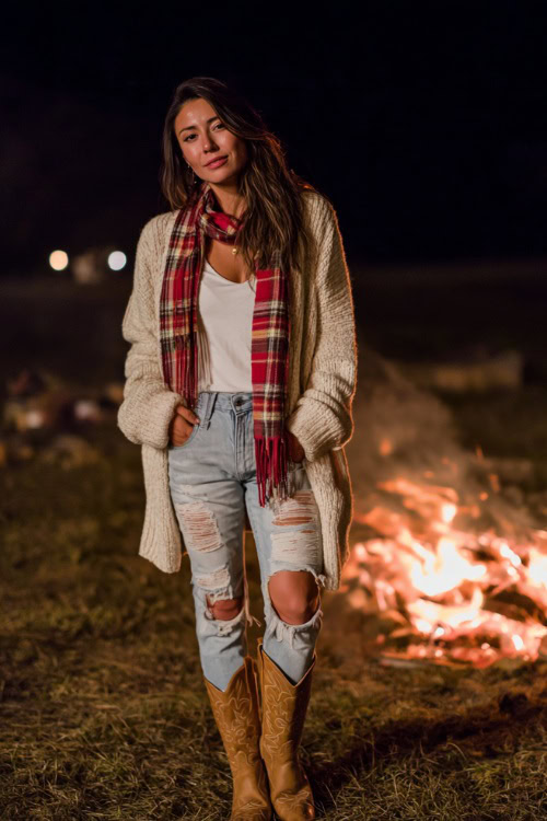 A woman wears light wash ripped jeans with a cozy oversized cardigan, styled with cowboy boots and a plaid scarf, standing by a bonfire at a country concert under fall night skies
