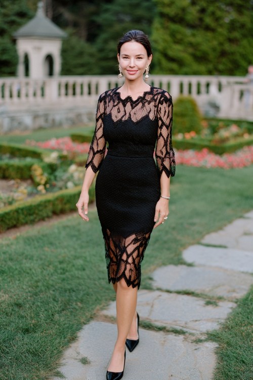A woman over 40 wears a black lace midi dress with sheer sleeves and scalloped hem