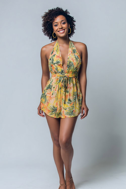 A Black woman wearing a halter mini dress in a tropical print with flat lace-up sandals and hoop earrings, styled for a carefree summer vibe, simple studio background