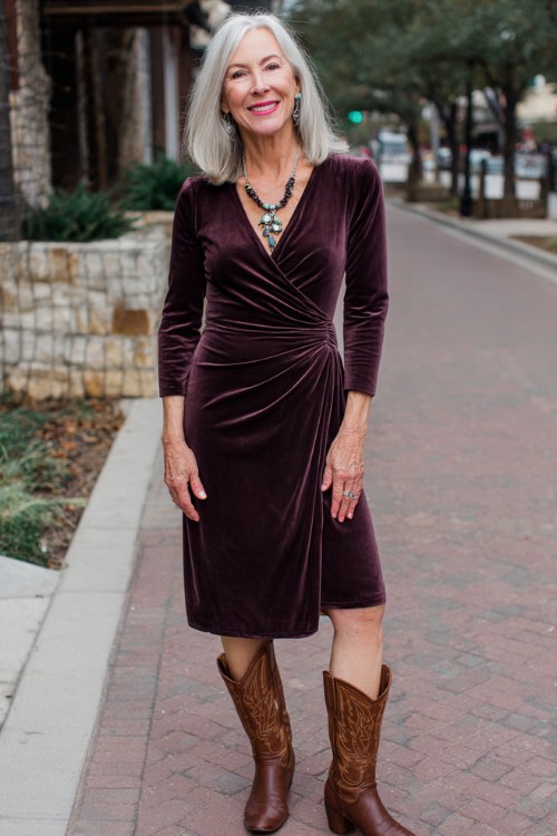 A woman over 50 wears a deep plum velvet midi dress with a faux-wrap design and long sleeves, paired with chocolate brown cowboy boots