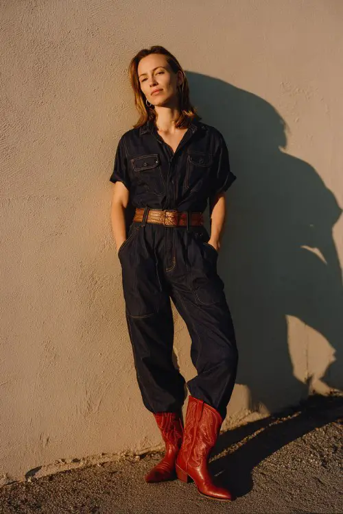 A woman wears red cowboy boots, a dark denim jumpsuit, and a brown leather belt