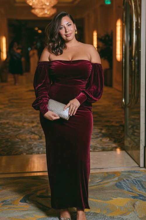 A curvy woman wears a burgundy velvet off-shoulder gown with sheer bishop sleeves