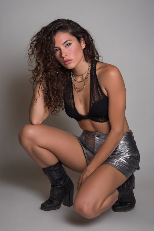 A woman in a metallic mini skirt, black sheer top with a bralette underneath, and ankle boots, styled for a bold summer night out, simple studio background