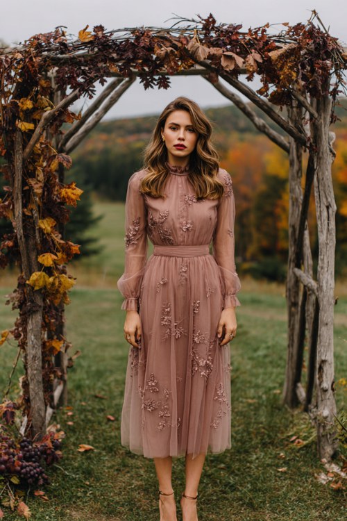 A woman wears a long-sleeve tulle midi dress in dusty rose with delicate floral embroidery, styled with heels