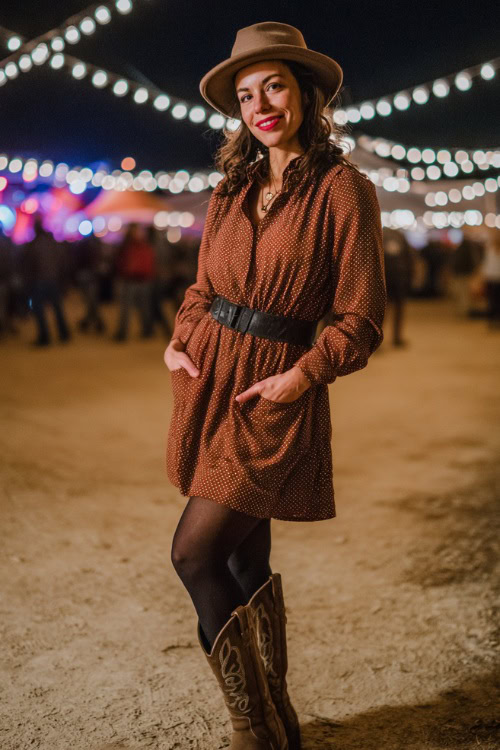 A woman over 40 wears a long sleeve knit sweater dress with a belted waist, black tights, and tall cowboy boots, styled with a wool fedora hat at a nighttime concert with string lights