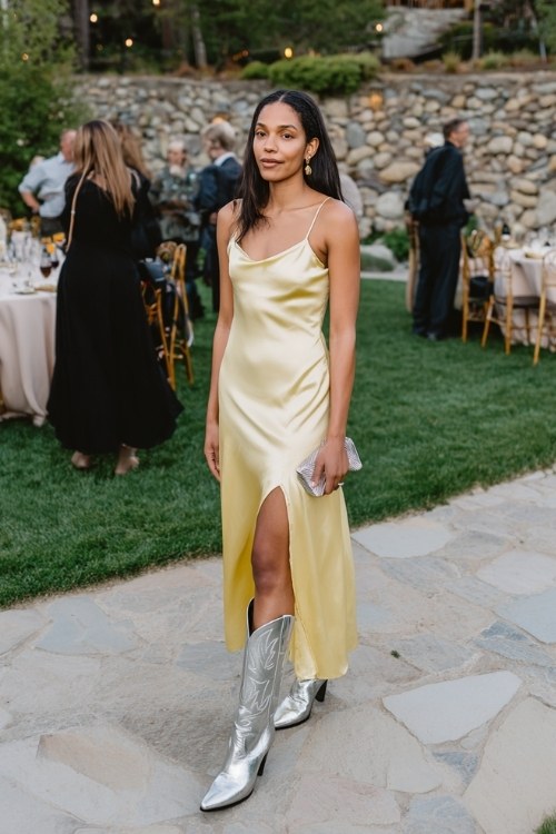 A woman over 30 wears a butter-yellow silk slip dress with a thigh slit, paired with metallic silver cowboy boots and a satin clutch