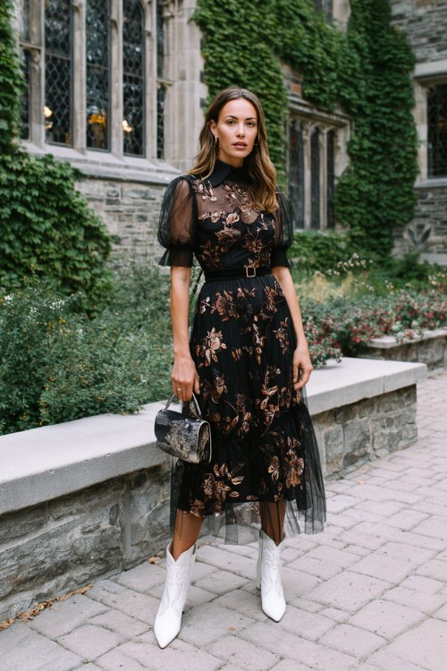 A woman over 30 wears a black tulle midi dress with embroidered copper florals, paired with white cowboy boots and a velvet clutch