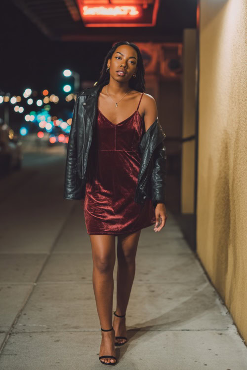 A Black woman in a burgundy velvet slip dress with a black leather moto jacket and ankle strap heels, walking under neon lights outside a night bar