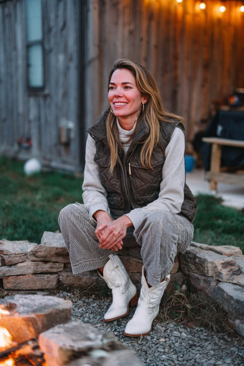 A woman over 30 wears wide-leg faded jeans, a quilted vest over a thermal top, and bright white cowboy boots