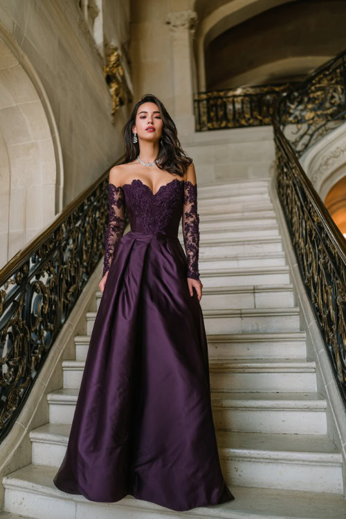 A woman wearing a deep purple floor-length gown with lace sleeves and silver jewelry, posing at the top of a grand staircase inside an elegant wedding venue