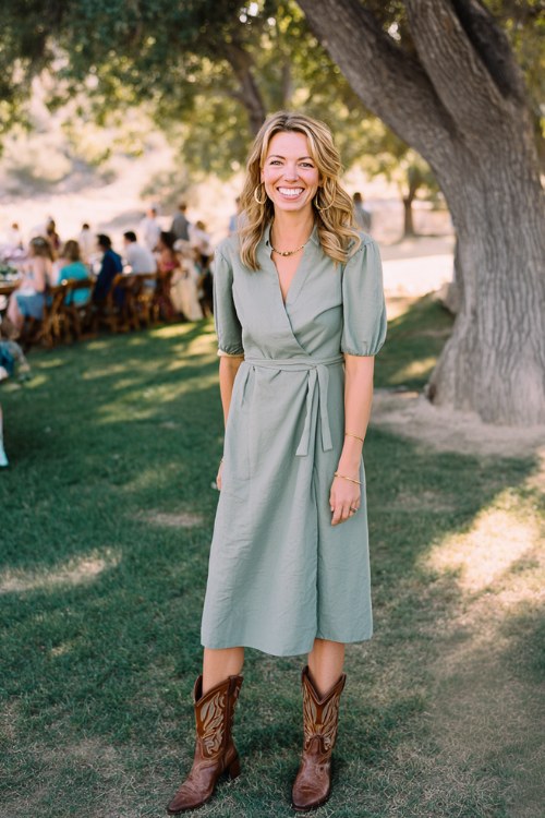 A woman over 30 wears a sage-green midi dress with brown cowboy boots and delicate gold jewelry