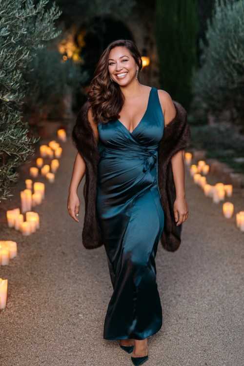 A curvy woman wears a dark teal satin gown with a wrap silhouette and faux-fur shawl