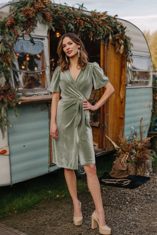 A woman wears a sage green velvet dress with flutter sleeves and wrap waist, styled with beige platform heels and drop earrings