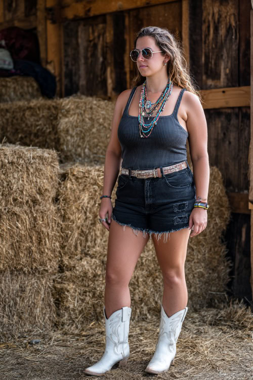 A plus-size woman in frayed black denim shorts, a sleeveless ribbed top, and white cowboy boots, accessorized with layered necklaces and aviator sunglasses