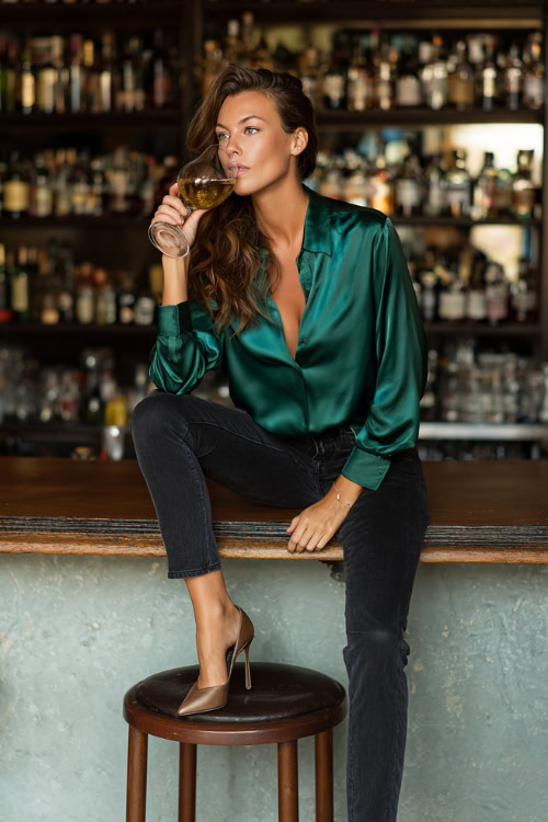 A woman in straight-leg dark jeans, a silky emerald blouse, and pointed-toe heels, sipping wine at a stylish bar counter