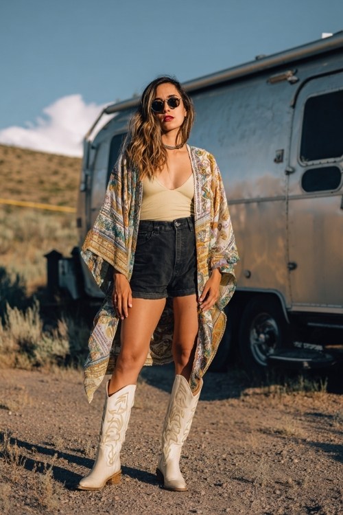 A woman over 30 wears black tailored shorts with cream tall cowboy boots and a flowy printed kimono