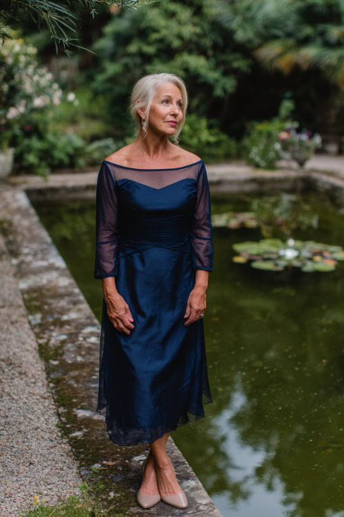 A woman over 60 wearing a navy blue midi dress with a boat neckline and sheer overlay, standing at the edge of a pond during a formal garden wedding