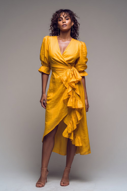 A Black woman wearing a bold yellow wrap dress with ruffle details and ankle-strap heels, styled for a radiant summer evening look, simple studio background