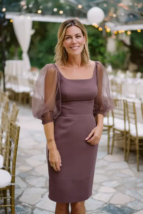 A woman over 30 wears a fitted dark mauve cocktail dress with three-quarter sheer sleeves