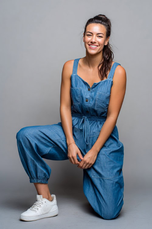 A woman in a sleeveless jumpsuit with a relaxed fit, cinched waist, and white sneakers, styled for casual outdoor dinner, simple studio background