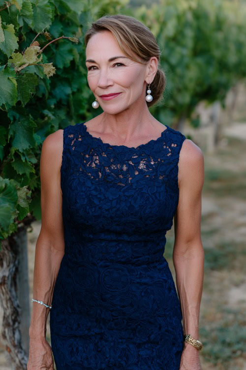 A woman over 60 wearing a navy blue knee-length sheath dress with lace overlay and pearl drop earrings, standing near a vineyard wedding setup