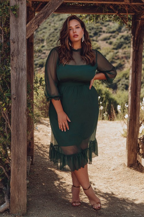A plus-size woman wears a forest-green chiffon midi dress with sheer sleeves and a ruffle hem, styled with ankle-strap heels and pearl earrings