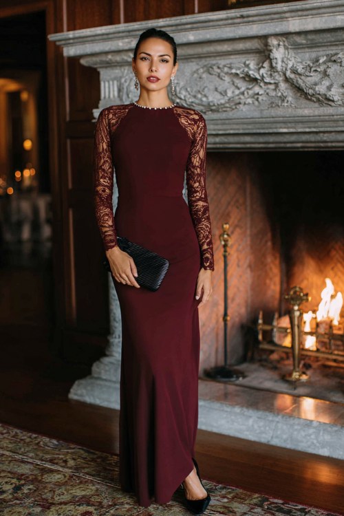 A woman wears a burgundy floor-length dress with lace sleeves, diamond studs, and black pumps
