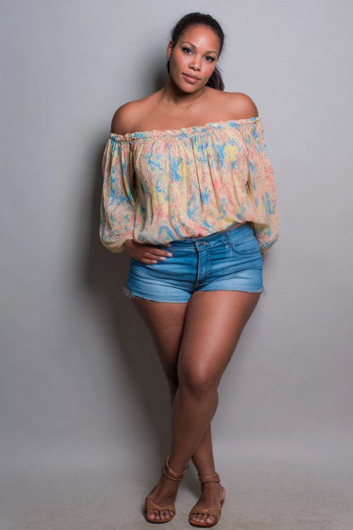 A plus-size woman in a flowy off-shoulder top paired with high-waisted denim shorts and flat sandals, styled for a casual summer night out, simple studio background