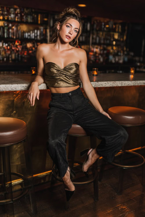 A woman wearing cropped black jeans, a metallic gold top, and block heels, posing at a nightclub bar with moody fall lighting, full body outfit visible