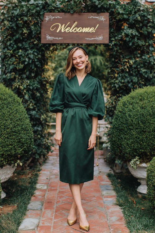 A petite woman wears a forest-green wrap midi with balloon sleeves and a waist sash, paired with gold slingback pumps and a velvet headband