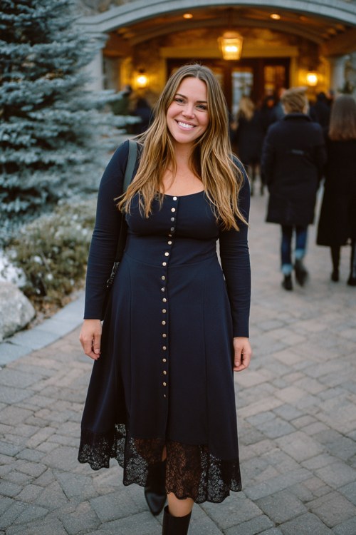 curvy woman wears a midnight blue long-sleeve dress with pearl button accents and a sheer lace hem, paired with dark boots and a longline wool coat
