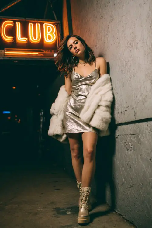 A woman wears a metallic mini dress layered with an oversized faux fur coat and platform boots, standing confidently under a glowing club sign as cold wind moves through