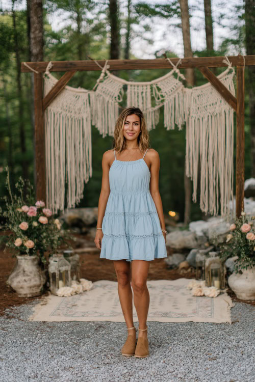 A woman in a floral sleeveless cotton midi dress with buttons down the front and flat sandals, standing under a rustic wooden arch covered in vines and flowers