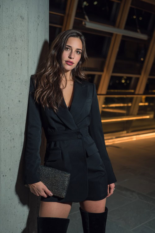 A woman wearing a tailored black blazer mini dress with long sleeves, paired with over-the-knee boots and a glittery clutch