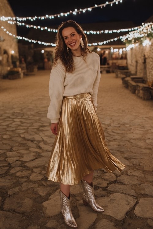 A woman wears a golden pleated midi skirt with a cream knit sweater tucked in, styled with silver cowboy boots and statement earrings