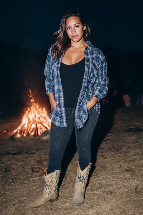 A curvy woman wears skinny jeans with an oversized navy flannel shirt as a jacket, layered over a fitted black tank, styled with embroidered cowboy boots