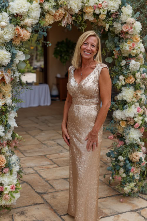 A woman over 50 in a champagne-colored sequin dress with cap sleeves and a V-neckline, standing under a wedding arch covered in summer florals