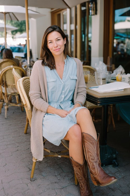 A woman wears a soft blue shirt dress with brown cowboy boots and a neutral cardigan