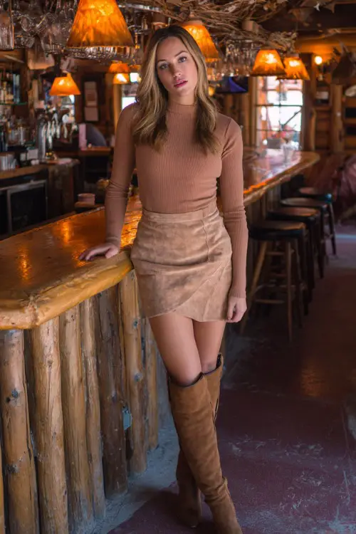 A woman in a tan suede skirt, a fitted long-sleeve top, and knee-high boots, standing inside a rustic bar with warm autumn lighting, full body outfit visible