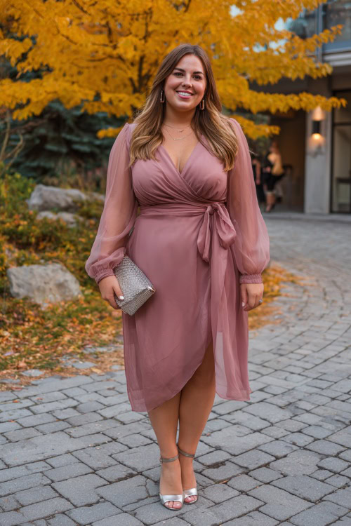 A plus-size woman wears a dusty rose wrap dress with long sheer sleeves and a tie waist, paired with silver block heels and a small clutch