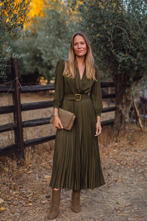 A woman wears a dark olive green pleated midi dress with a belted waist, paired with suede ankle boots and a clutch