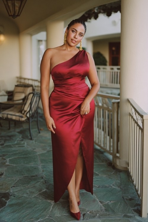A curvy woman wears a deep red satin gown with a one-shoulder draped detail